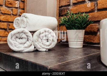 Ecke des Badezimmers auf einer grauen Oberfläche weiße Handtücher sind in Rollen Loft-Stil Ziegelwände grün Topfgras gestapelt. Stockfoto