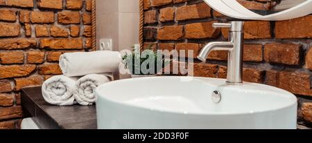 Badezimmer weißes Waschbecken auf der grauen Oberfläche der gestapelten Rollen von weißen Handtüchern Loft-Stil Ziegelwand grünes Gras im Topf Banner. Stockfoto