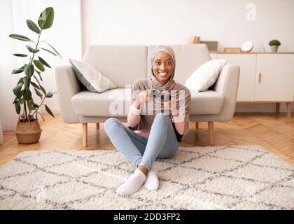 Happy Muslim Frau in Hijab sitzen auf dem Boden mit Joystick, Videospiel zu Hause spielen, in voller Länge Porträt Stockfoto