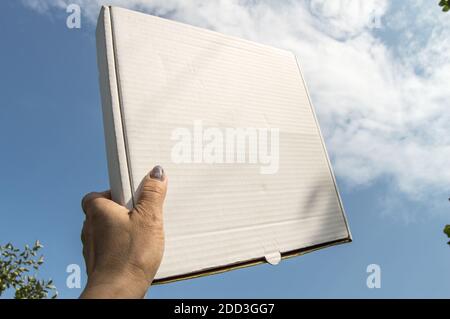 Eine Frauenhand hält eine weiße Pappschachtel mit einer Kopie des Raumes und des Platzes für Text auf einem blauen Himmel Hintergrund, Schablone, mockup Stockfoto