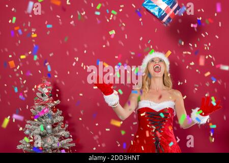 Schöne junge Blondine in Weihnachtsmann Kleid, zwischen fallenden Konfetti mit offenen Armen zu sammeln Geschenk, das vom Himmel fällt, neben dem verschneiten und de Stockfoto