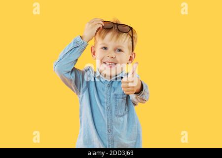 Portrait von überrascht niedlichen kleinen Jungen in Sonnenbrille. Kind mit Spaß isoliert über gelben Hintergrund. Blick auf die Kamera. Daumen hoch zeigen Stockfoto