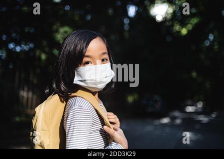 Portrait von kleinen japanischen Mädchen mit Rucksack zu Fuß im Freien in der Stadt, Coronavirus Konzept. Stockfoto