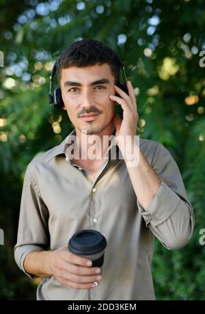 Junger Mann mit Kopfhörer und Kaffee in wiederverwendbarer Tasse im Freien in der Stadt stehen. Stockfoto