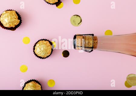 Flasche rosa Champagner mit Konfetti, goldene Süßigkeiten auf rosa Hintergrund. Festliche Pastellgold Feier flach legen Stockfoto