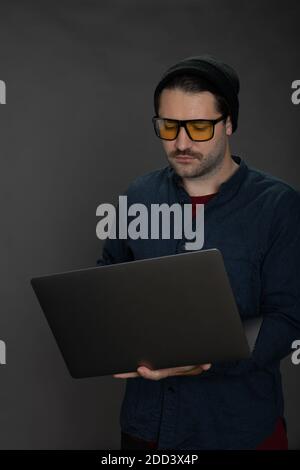 Schöner unrasiert Hipster in gestrickten Mütze arbeiten mit Laptop an Grauer Hintergrund Stockfoto