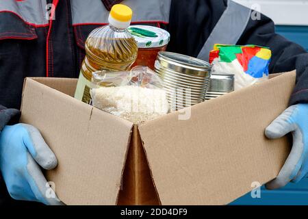 Ein Freiwilliger in einer Winterjacke hält einen Karton mit trockenem Getreide, Konserven, Butter und anderen Produkten. Hilfe während einer Pandemie. Spenden von fo Stockfoto
