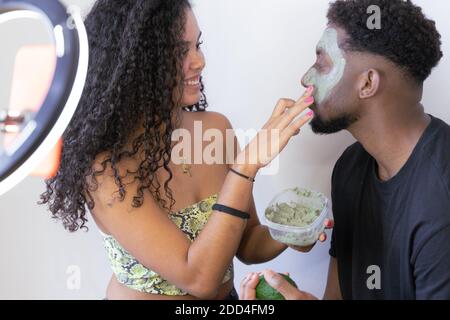 Frau, die Avocado Maske auf das Gesicht ihres Freundes. Junge liebevolle Paar kümmert sich um die Haut zu Hause. Influencer koppeln Videoaufnahme. Stockfoto