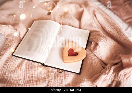 Leckerer herzförmiger Keks mit Erdbeermarmelade auf offenem Papierbuch über glühenden Lichtern aus nächster Nähe. Valentinstag. Stockfoto