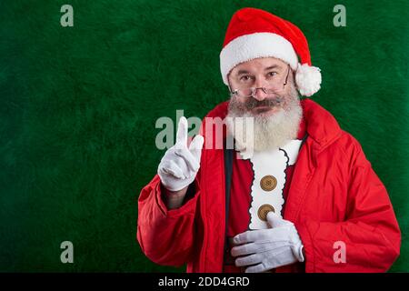 Reifer bärtiger Mann mit Brillen im Gesicht trägt Weihnachtsmütze, erhobener Zeigefinger, Aufmerksamkeitsschild, Kopierraum Stockfoto