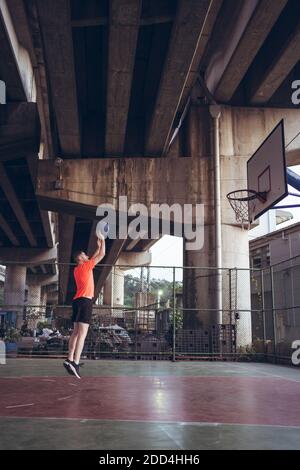 Kaukasischer junger Mann spielt Basketball allein auf städtischen Strecke unter highway Bridge in orangefarbenem T-Shirt Stockfoto