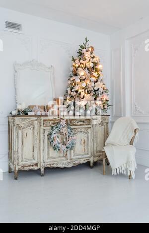 Geschmückter Weihnachtsbaum auf hölzerne Kommode Kommode Kommode in weiße Interieur mit künstlichen Blumen, Girlanden und Spielzeug Stockfoto