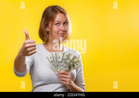 Finanzen, Darlehen und Banken. Eine junge Blondine hält einen Geldwad in ihren Händen und gibt einen Daumen nach oben, lächelnd. Gelber Hintergrund. Speicherplatz kopieren. Stockfoto
