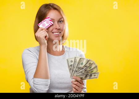 Finanz- und Bankkarten. Eine junge Blondine hält einen Geldwad in ihren Händen und bedeckt lächelnd ihre Augen mit einer Bankkarte. Gelber Hintergrund und Kopie Stockfoto