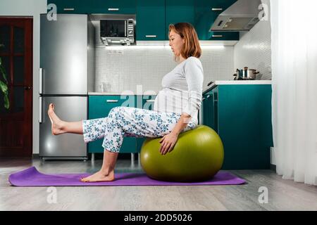 Selbstisolierung und Heimtraining. Eine schwangere kaukasische Frau sitzt auf einem Fitball, lehnt sich mit den Händen darauf und hebt die Beine. Küche im Th Stockfoto