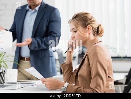 Verärgerte Geschäftsfrau mit Stift, der Dokument ansieht, während sie am Arbeitsplatz mit verschwommenem Kollegen auf dem Hintergrund sitzt Stockfoto