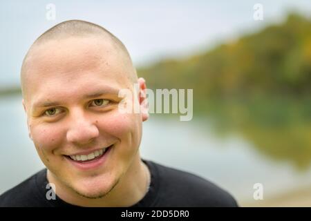 Unrasiert junger Mann mit einem schönen warmen freundlichen Lächeln in Ein Nahaufnahme Kopf Porträt Blick auf die Seite von Der Rahmen mit ruhigen See Hintergrund Stockfoto
