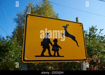 Ein gelbes Straßenwarnschild mit einer lustigen Gegenüberstellung von Schulkindern und springenden Hirschen, die grafische Bilder kreuzen. Auf Deer Isle, Maine. Stockfoto