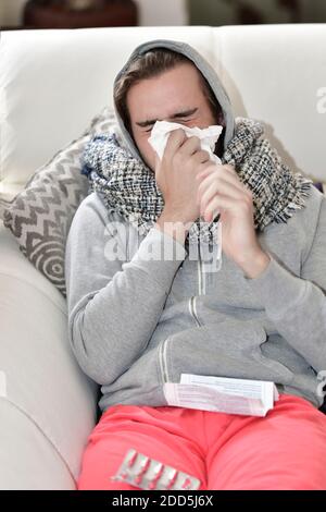 Blick auf einen jungen Mann mit Kapuzenpullover und einem Schal, der seine Nase mit einer Papierserviette auf einem unscharf Hintergrund bläst. Krankheit und Medizin Konzept. Stockfoto