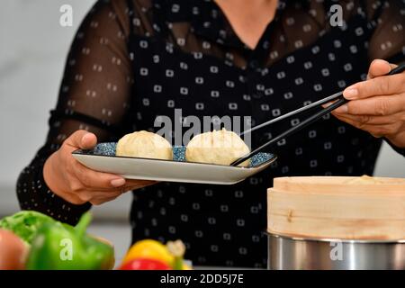 Nahaufnahme einer Frau, die einen Teller mit zwei verführerischen Knödeln hält, die einen mit Essstäbchen aufsammeln. Traditionelles chinesisches Essen und Kochkonzept. Stockfoto