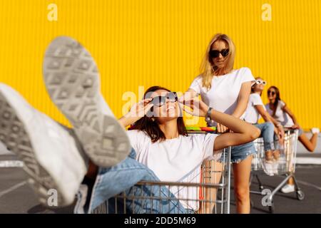 Zwei schöne glückliche junge Frauen, Freundinnen in weißen T-Shirts haben Spaß auf Einkaufswagen, auf dem Hintergrund eines Einkaufszentrums auf einer sommerlichen sonnigen da Stockfoto
