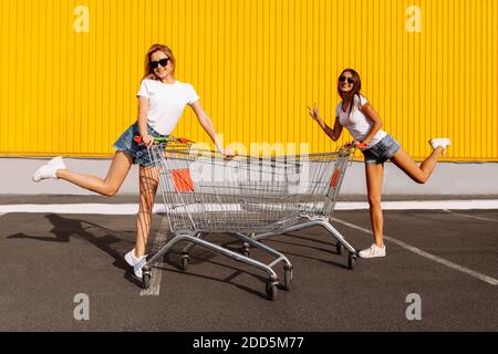 Zwei schöne glückliche junge Frauen, Freundinnen in weißen T-Shirts haben Spaß auf Einkaufswagen, auf dem Hintergrund eines Einkaufszentrums auf einer sommerlichen sonnigen da Stockfoto