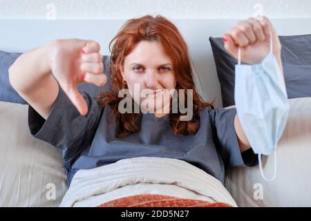 Rothaarige Frau nahm die Maske ab und zeigt einen Finger nach unten. Frau mit Coronavirus zu Hause im Bett Stockfoto