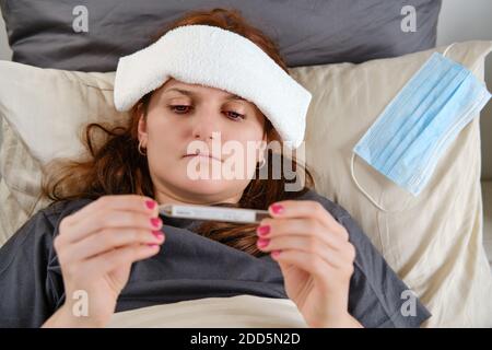 Frau mit roten Haaren liegt im Bett krank mit Coronavirus Mit einer kalten Kompresse auf dem Kopf Stockfoto