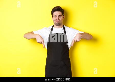 Zweifelhafter Barista, der sich beschwert, mit den Fingern nach unten zeigt und unzufrieden grimaziert, steht in schwarzer Schürze vor gelbem Hintergrund Stockfoto
