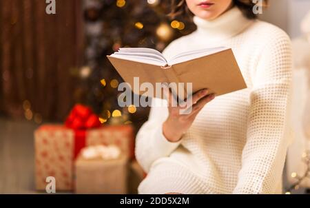 Ein Mädchen in einem weißen Pullover sitzt und liest ein Buch auf dem Hintergrund eines Weihnachtsbaums und Geschenke Stockfoto