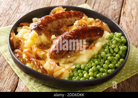 Schmackhafte Bangers und Mash aus Würsten serviert mit Kartoffelpüree, grünen Erbsen und Zwiebelsoße in Nahaufnahme auf einem Teller auf dem Tisch. Horizontal Stockfoto