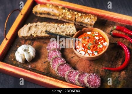 Rustikales Holztablett mit einfachen Balkan-Lebensmitteln - Ajvar Aufstrich, Brot, Fleisch, Knoblauch und Chili (selektiver Fokus) Stockfoto