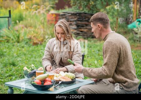 Ein junges Paar bereitet sich auf ein Familien-Barbecue-Dinner im Innenhof des Hauses vor. Gemüse auf dem Picknicktisch schneiden. Ein warmer Abend in den letzten Sommertagen Stockfoto