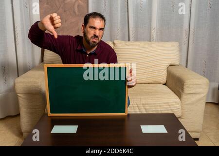 Bärtiger Mann, der zu Hause mit dem Daumen nach unten auf der Couch sitzt. Konzept der Hoffnungslosigkeit Stockfoto