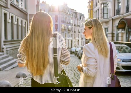 Zwei junge Frauen mit Laptoptaschen gehen entlang der Straße der Sunset City, Rückansicht Stockfoto