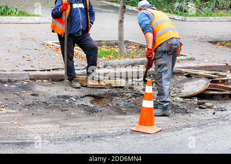 Straßenarbeiter, die in reflektierender Kleidung gekleidet sind, benutzen eine Brechstange und eine Schaufel, um ein altes Schachtloch auf einem eingezäunten Straßenabschnitt zu demontieren. Stockfoto