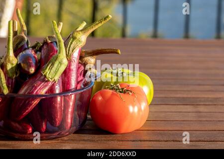 Konzentrieren Sie sich auf das Sonnenlicht, das auf eine rote Tomate reflektiert. Auf dem hölzernen Tisch eine Gruppe von kleinen Auberginen im Glas und eine andere grüne Frucht. Hintergrund für das Essen Stockfoto