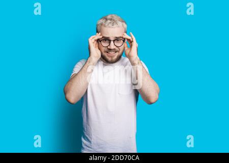 Überrascht kaukasischen Mann mit Bart und blonde Haare berühren seine Head and Wear Brillen posiert auf einem blauen Studio Hintergrund Stockfoto