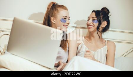 Kaukasische Freunde mit Anti-Falten-Masken unter den Augen lachend in Bett mit Laptop Stockfoto