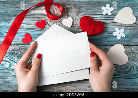 Eine Frau hält einen weißen Umschlag mit einem leeren Brief auf einem romantischen Hintergrund mit Herzen und einem Band. Stockfoto