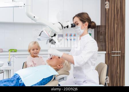 Eine blonde Frau, die ihre Zähne im Zahnarztbüro überprüft hat, und ihre Tochter wartet auf sie. Stockfoto