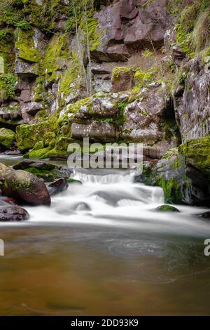 Nahaufnahme einer kleinen Kaskade auf einem Gebirgsfluss, der durch die enge, rote Felsenschlucht mit Felsen fließt, die von grünem, herbstfarbenem Moos bedeckt sind Stockfoto