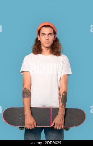 Selbstbewusster junger erwachsener Mann in trendigen weißen T-Shirt und orangefarbenem Hut, im Studio auf blauem Hintergrund stehend, Skateboard in den Händen haltend Stockfoto