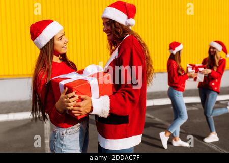 Fröhliche schöne junge Frauen in weihnachtspullover und weihnachtsmann Hüte feiern weihnachten und schenken einander Geschenke gegen Gelb Wand im Freien Stockfoto