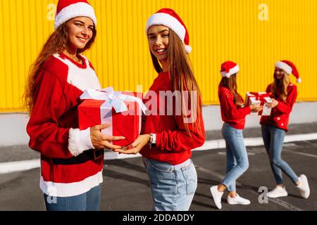 Fröhliche schöne junge Frauen in weihnachtspullover und weihnachtsmann Hüte feiern weihnachten und schenken einander Geschenke gegen Gelb Wand im Freien Stockfoto