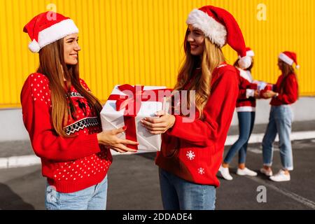 Fröhliche schöne junge Frauen in Weihnachtspullover und Weihnachtsmann Hüte feiern Weihnachten und schenken einander Geschenke im Freien Stockfoto