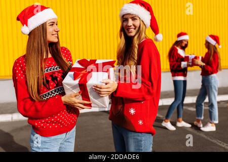 Fröhliche schöne junge Frauen in weihnachtspullover und weihnachtsmann Hüte feiern weihnachten und schenken einander Geschenke gegen Gelb Wand im Freien Stockfoto