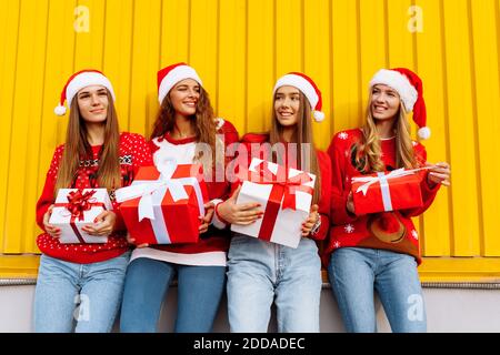 Glücklich aufgeregt Gruppe von Freunden in roten Pullover gekleidet und weihnachtsmann Hüte halten weihnachtsgeschenke, während in der Nähe gelb stehen Wand in der Nähe Stockfoto