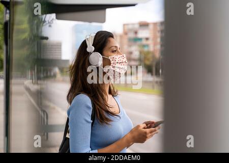 Betrachten Frau in Gesichtsmaske tragen Kopfhörer mit Smartphone Am Busbahnhof während der COVID-19 Stockfoto