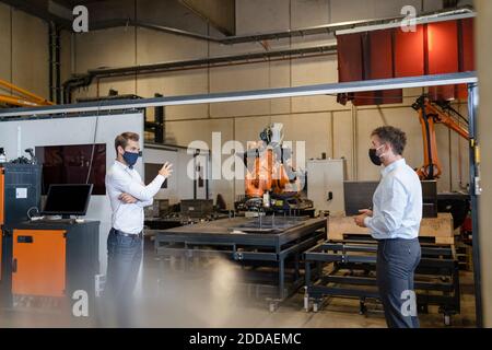 Geschäftsleute mit Gesichtsmaske diskutieren, während sie in sozialer Distanz stehen Gegen Roboterarmgeräte im Werk Stockfoto